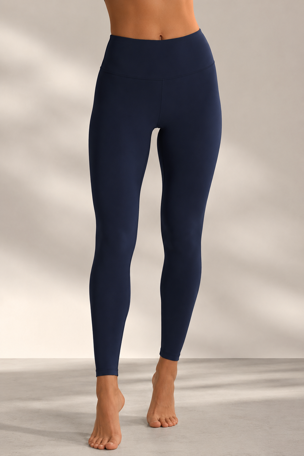 A model from the waist down wearing navy blue high-waisted leggings, standing barefoot against a neutral studio background with soft shadows.