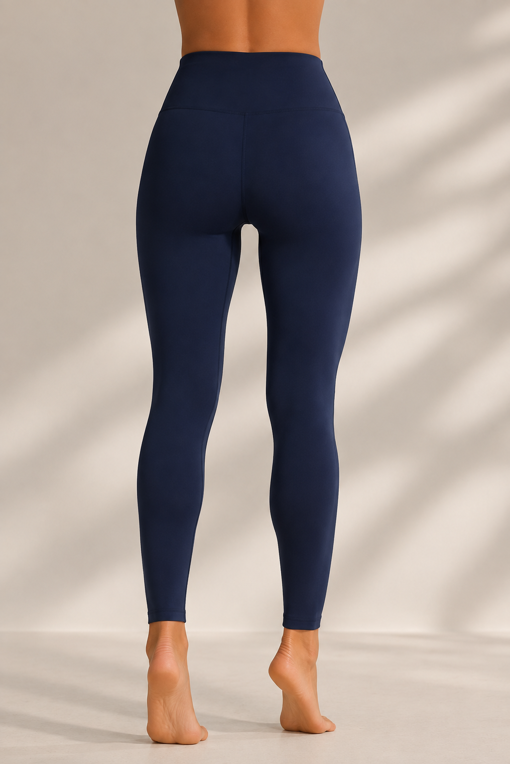 A person shown from the back wearing high-waisted navy blue leggings, standing barefoot against a warm beige wall with soft diagonal shadows.